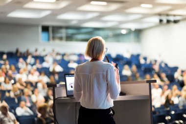 Female speaker giving a talk on corporate business conference. Unrecognizable people in audience at conference hall. Business and Entrepreneurship event clipart