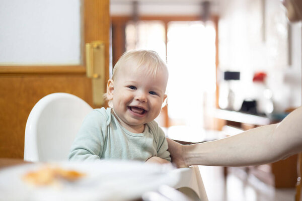 Adorable cheerful happy infant baby boy child smiling while sitting in high chair at the dining table in kitchen at home beeing spoon fed by his mother.