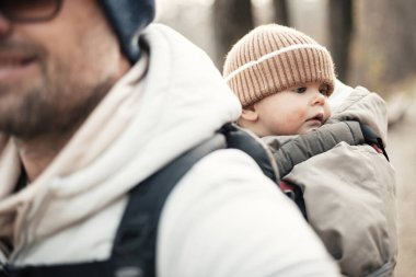 Yavru oğlunu taşıyan sportif baba sonbahar ormanlarında yürüyüş yaparken sırt çantasında tulum ve şapka giyiyor.