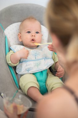 Anne, bebeğini bebek koltuğunda meyve püresiyle besliyor. Bebek katı gıda tanıtım konsepti