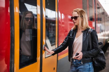 Kot pantolon, gömlek ve deri ceket giyen, güneş gözlüğü takan genç sarışın kadın tren istasyonunda trene binmek için modern hız treninin kapı tuşuna basıyor. Seyahat ve ulaşım.