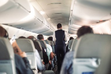 Uçakta koltuklarda yolcular ve koridorda yürüyen, insanlara hizmet eden üniformalı hostesler var. Ticari ekonomi uçuş hizmeti kavramı
