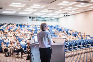 Female speaker giving a talk on corporate business conference. Unrecognizable people in audience at conference hall. Business and Entrepreneurship event clipart