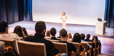 Business and entrepreneurship symposium. Female speaker giving a talk at business meeting. Audience in conference hall. Rear view of unrecognized participant in audience. clipart