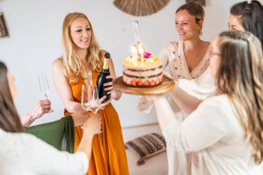 Doğum günü sürpriz partisi. Bir grup neşeli bayan arkadaş kişisel başarı hikayelerini kutluyorlar. Şampanyayla kadeh kaldıran gülümseyen kızlar. Biri elinde mumlarla doğum günü pastası tutan kızlar..