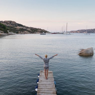 Tahta iskele kollarında duran güzel kadın alacakaranlıkta huzurlu deniz manzarasının keyfini çıkarıyordu. Dişi gezgin Porto Rafael, Costa Smeralda, Sardinya, İtalya 'da tahta bir iskelede duruyor..