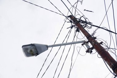 Sokak lambası bulutlu bir gökyüzüne karşı duruyor, paslı direği elektrik kablolarına dolanmış. Kentsel bozulma ve altyapı zorluklarını çağrıştırıyor.