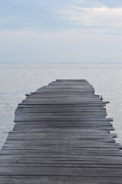 A weathered wooden pier extends into calm ocean waters beneath a pale, expansive sky, evoking a sense of peace and invitation for escape or reflection.