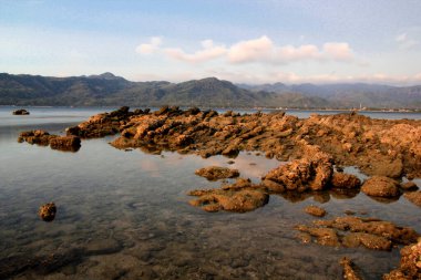 Explore a serene coastal landscape with unique rock formations and calm waters, reflecting distant green mountains and a soft sky.