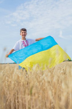 Ukrayna için dua et. Buğday tarlasında Ukrayna bayrağı taşıyan bir çocuk. Ulusal Ukrayna elbiseli gülümseyen çocuk elinde sarı-mavi büyük bayrak ile buğday tarlasının arka planında. Vatansever eğitim.