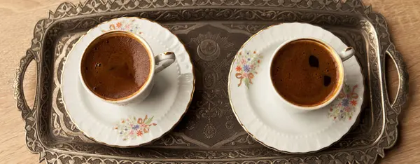 Traditional foamy Turkish coffee in a patterned white cup in a metal tray on an oak coffee table