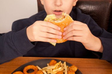 Beyaz bir adam yarısı yenmiş hamburger, patates cipsi, soğan halkası ve tavuk parçalarını ahşap bir masadaki gri tabaktan, Amerikan fast food kültüründen elde yer.