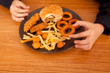 Beyaz bir adam yarısı yenmiş hamburger, patates cipsi, soğan halkası ve tavuk parçalarını ahşap bir masadaki gri tabaktan, Amerikan fast food kültüründen elde yer.