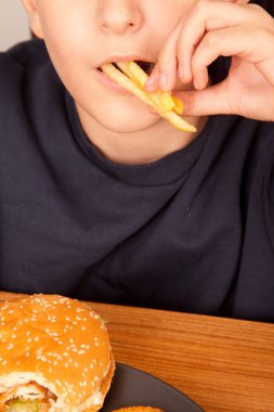 Beyaz bir adam yarısı yenmiş hamburger menüsü yer. Amerikan fast food kültürü olan ahşap bir masada gri bir tabaktan elde patates cipsi.