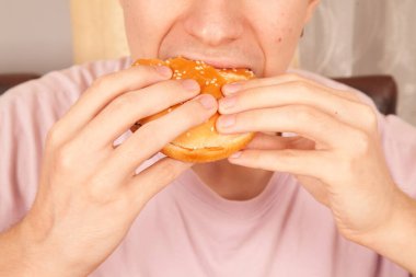 Beyaz bir adam yarısı yenmiş susamlı hamburger yiyor. Amerikan fast food kültürü.