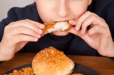 Beyaz adam kızarmış tavuk kanadı, hamburger, patates cipsi, soğan halkası ve ahşap masada gri tabakta tavuk kanadı parçaları yiyor. Amerikan fast food kültürü.