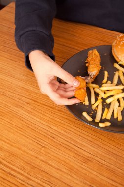 Beyaz adam kızarmış tavuk kanadı, hamburger, patates cipsi, soğan halkası ve ahşap masada gri tabakta tavuk kanadı parçaları yiyor. Amerikan fast food kültürü.
