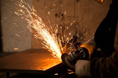 Ağır bir sanayide ya da fabrikada çalışan bir işçi elinde kesme taşı ya da öğütme taşı tutuyor ve metalden kıvılcımlar çıkıyor.