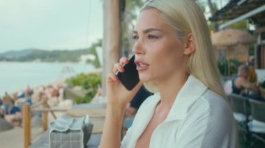 Beyaz tişörtlü sarışın bir kafede telefonla konuşuyor.
