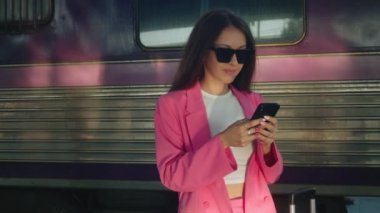 A gorgeous woman in a pink suit is standing at the train station talking on her mobile phone. Looks stylish and charming