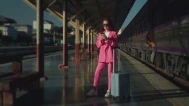 A gorgeous woman in a pink suit is standing at the train station talking on her mobile phone. Looks stylish and charming