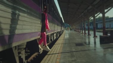 A young woman in a pink suit gets off the train on the platform and walks toward the camera in slow motion