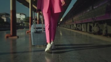 A young woman in a pink suit and with a suitcase walking on the platform near the train in slow motion, close-up follow shot