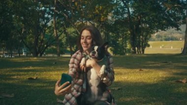 Young woman takes selfie with her Boston Terrier at sunset in park in slow motion