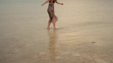 Güzel, uzun saçlı kız gün batımında kumsalda ağır çekimde yürüyor. Anamorfik çekim