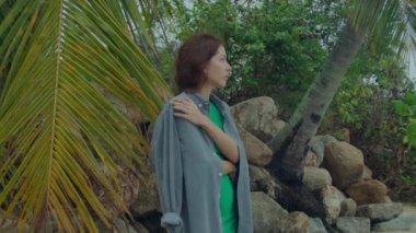 Genç bir kadın bulutlu bir deniz sahilinde tek başına duruyor, üzgün bir şekilde ufka bakıyor.