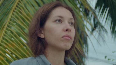 Genç bir kadın bulutlu bir deniz sahilinde tek başına duruyor, üzgün bir şekilde ufka bakıyor.