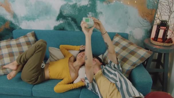 レトロなスタイルのカメラでスナップショットを撮るのが大好きな若いカップル ゆっくりとした動きで彼らの部屋のソファの上で彼らの自由な時間で笑い リラックス — ストック動画