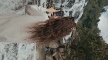 Genç bir kadın Kuzey Tayland 'da görkemli bir şelalenin fotoğrafını çekiyor. Yavaş çekim dikey video