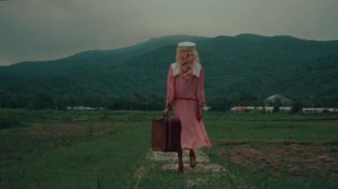 Antika elbiseli ve eski bir bavul taşıyan genç bir kız bir dağın önünde küçük bir köprüden geçiyor. Yüksek kalite 4 bin yavaş çekim görüntüsü