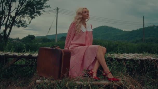 背景に山のある橋の上に座っているヴィンテージのドレスとレトロなスーツケースの女性 高品質4Kスローモーション映像 — ストック動画
