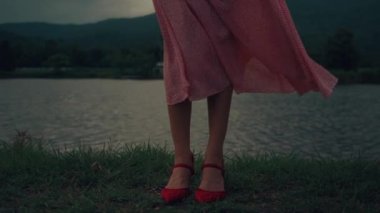 Kırmızı elbiseli ve kırmızı ayakkabılı bir kadın gölün kenarında duruyor, elbisesi rüzgarda sallanıyor. Yüksek kalite ağır çekim 4k görüntü