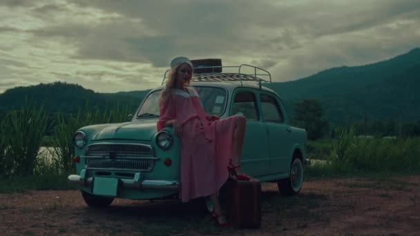 一个穿着粉色复古服装的女孩坐在青绿色复古车的车篷上 背靠山脉背景的肖像 高质量的4K慢镜头 — 图库视频影像