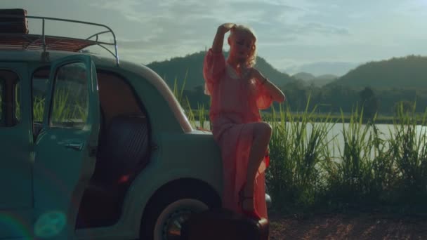 一个穿着粉色复古服装的女孩坐在一辆深蓝色复古轿车的行李箱上 背靠山脉背景的肖像 高质量的4K慢镜头 — 图库视频影像