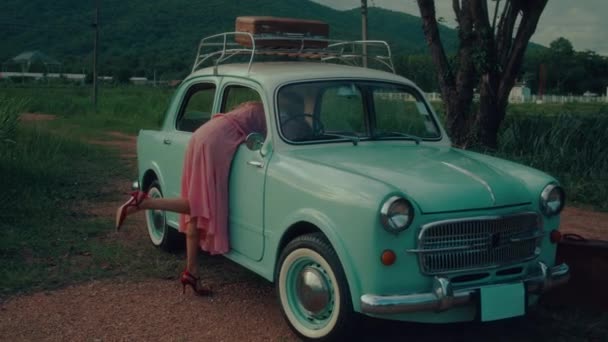 山の背景に対してレトロ車のドアに掛かるヴィンテージピンクのドレスの長髪の少女 — ストック動画