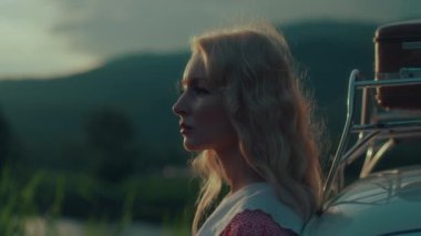 Uzun saçlı sarışın gün batımında gölün yanındaki dağlarda eski arabasının üzerinde duruyor.