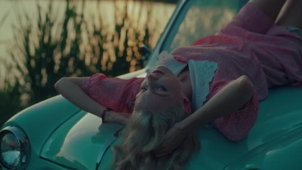 日落时 一位穿着粉色复古礼服的金发姑娘躺在一辆蓝色复古汽车的车篷上 高质量的4K慢镜头 — 图库视频影像