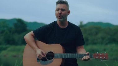 Gitarist göl kenarında çalıyor ve şarkı söylüyor. Yüksek kaliteli el kamerası görüntüsü 4K