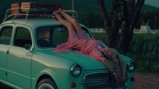 日落时 一位穿着粉色复古礼服的金发姑娘躺在一辆蓝色复古汽车的车篷上 高质量的4K慢镜头 — 图库视频影像
