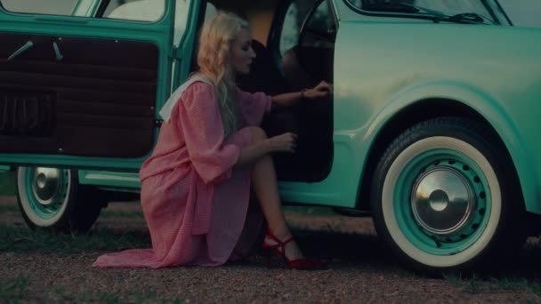 一个穿着粉色复古服装的女孩坐在一辆深蓝色复古轿车旁边的肖像 高质量的4K慢镜头 — 图库视频影像