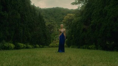 Lüks mavi elbiseli çekici kadın selvi ağaçlarıyla dolu bir sokakta yürüyor. Selvi ağaçlarında poz veren zarif bir kız. Ormanda manken olmak. Kara büyü 6k görüntüsü