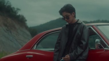 Kahverengi deri ceketli vahşi bir erkek fatma kız klasik kırmızı arabasına yaslanıp sigara içiyor. Kara büyü 6k görüntüsü