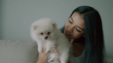 Genç Taylandlı bir kadın kucağında küçük şirin beyaz bir köpek, oturma odasında koltuğunda oturuyor, uzun saçlı genç bir kadın küçük tüylü bir köpek tutuyor ve gülümsüyor. İnsanlar ve hayvanlar arkadaşlık kavramı. Yüksek