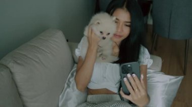 Genç Taylandlı bir kadın tatlı beyaz köpeğiyle selfie çekiyor oturma odasındaki kanepesinde uzanmış modern bir iç mekan ile. Esmer kadın küçük bir köpeğe sarılıyor ve akıllı telefonuyla fotoğraf çekiyor. İnsanlar