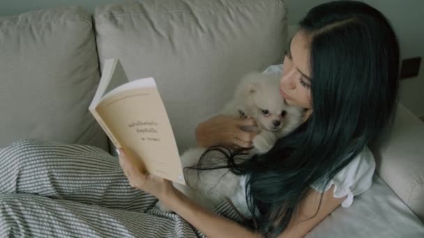 Młoda Czarnowłosa Kobieta Czyta Książkę Uroczym Białym Pluszowym Psem Leżącym — Wideo stockowe