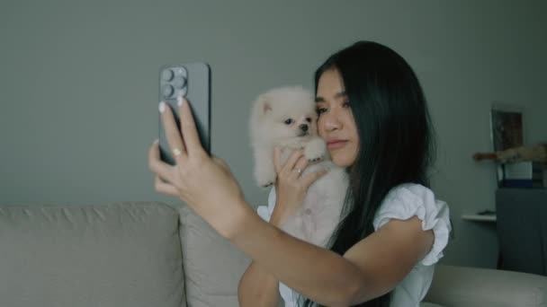 年轻的泰国人女人带着一只可爱的白色飞溅犬 躺在客厅的沙发上 房间里有一个现代的内饰 黑发女人抱着一条小狗 用她的智能手机自画像 — 图库视频影像